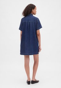 Jeansklänning med korta ärmar, mörkblå färg och avslappnad passform. Har en bakre infällning och fransig nederkant. Bärs med svarta ballerinaskor.