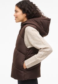 Smeđa podstavljena prsluk s kapuljačom, patentnim zatvaračem, bočnim džepovima i rebrastim rukavima džempera. Glatka tekstura s prošivenim dijelovima.