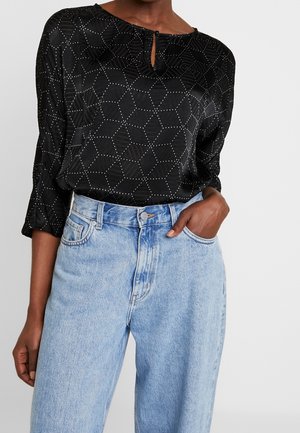 Sort mønstret bluse med lange ærmer og nøglehulshals, stoppet ned i højttaljede lyseblå jeans, den ene hånd i lommen.