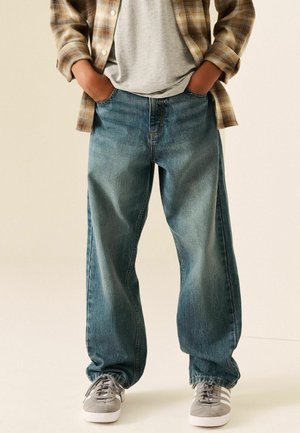 Persona che indossa jeans blu larghi, sneakers grigie, t-shirt grigia e camicia a quadri marrone con le mani nelle tasche.