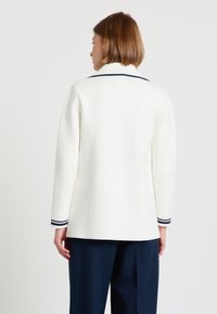 Frau mit kurzen braunen Haaren, die einen weißen Pullover mit langen Ärmeln und schwarzen Streifen an Bündchen und Kragen trägt, kombiniert mit marineblauen Hosen, mit dem Rücken zur Kamera.