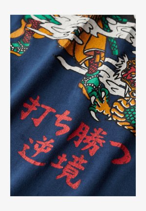 Mørkeblå stof med et farverigt grafisk design af en drage og en tiger, med rød japansk tekst i bunden.