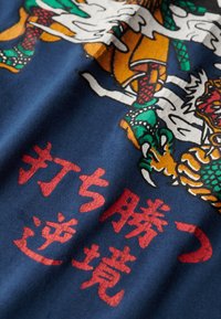 Le tissu bleu marine présente un graphisme détaillé d'un dragon et un texte stylisé en rouge. Le dragon a des écailles vertes et des accents orange.