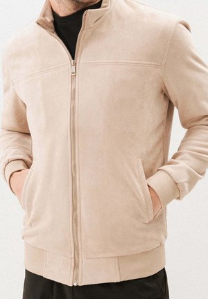 Man wearing beige zip-up jacket with hands in side pockets over black shirt and black pants, shown from neck to hips.