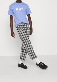 Světle modré bavlněné tričko s nápisem "HUF" vytištěným bílou barvou. Černobílé kárované kalhoty, lesklé černé mokasíny a bílé ponožky.