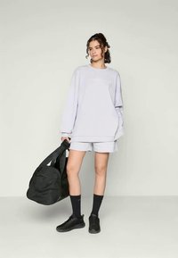 Light gray oversized sweatshirt with logo, paired with matching shorts. Black athletic shoes and a large black duffel bag. Plain background.