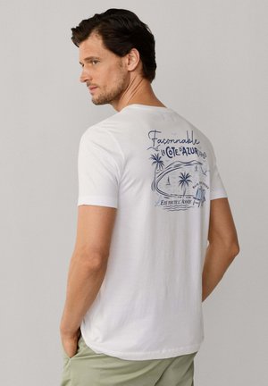 Mann in weißem T-Shirt mit blauem Küstendesign und französischem Text auf dem Rücken, seitlich stehend mit Händen in den Taschen vor einfachem Hintergrund.
