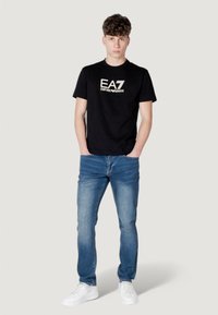 Camiseta de algodón negra con el logo "EA7 Emporio Armani", combinada con vaqueros de mezclilla azules y zapatillas blancas. Hombre de pie con las manos en los bolsillos.