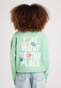 Felpa verde menta con testo bianco: "QUESTO DEVE ESSERE IL POSTO" e toppe colorate. Collo a giro, maniche lunghe e vestibilità rilassata.
