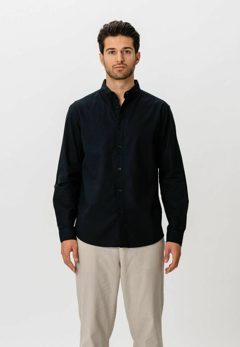 Homme aux cheveux foncés portant une chemise noire à boutons et un pantalon beige, debout devant un fond blanc uni, faisant face à l'avant.
