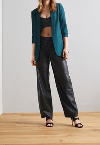 Turkusowa marynarka o dopasowanej sylwetce, zestawiona z czarnym krótkim topem i czarnymi spodniami typu wide-leg z sztucznej skóry, uzupełniona o czarne sandały na obcasie.