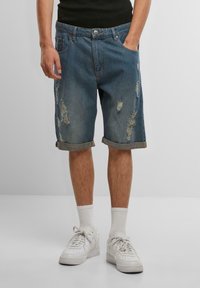 Shorts en denim bleu moyen avec des détails effilochés, des ourlets roulés et une coupe décontractée. Associés à des baskets blanches et des chaussettes de longueur mi-mollet.