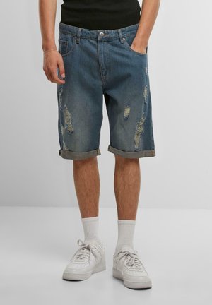 Shorts en denim bleu moyen avec des détails effilochés, des ourlets roulés et une coupe décontractée. Associés à des baskets blanches et des chaussettes de longueur mi-mollet.
