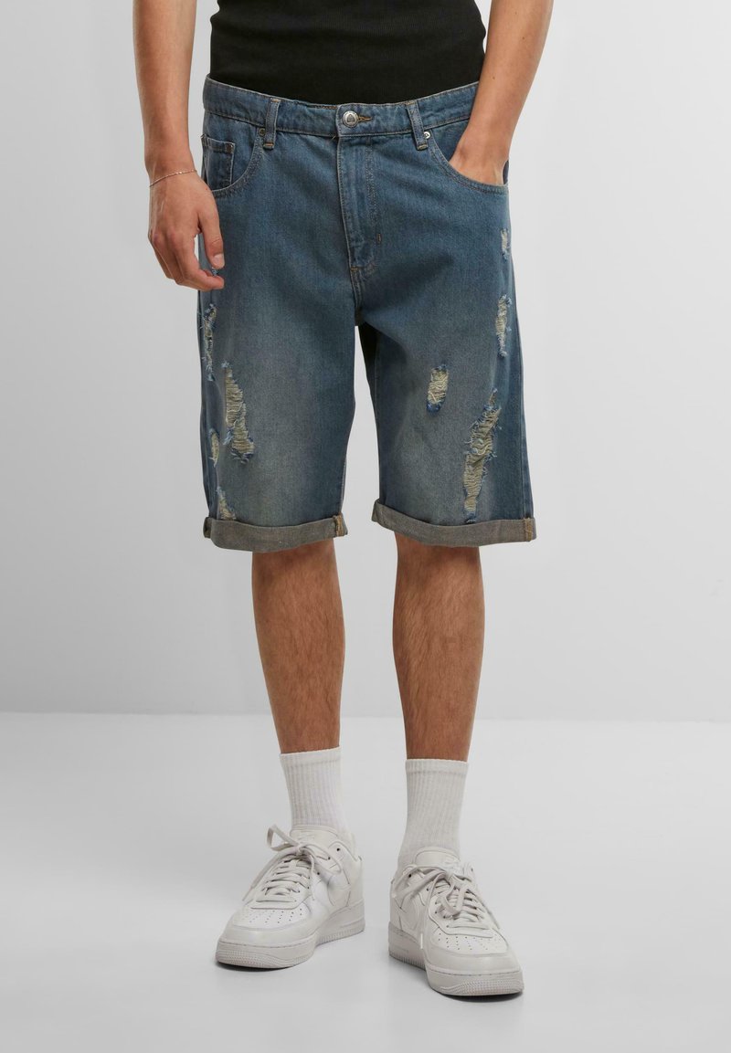 Shorts en denim bleu moyen avec des détails effilochés, des ourlets roulés et une coupe décontractée. Associés à des baskets blanches et des chaussettes de longueur mi-mollet.