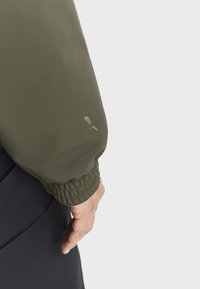 Olivgrüne Jacke mit elastischen Bündchen, die ein dezentes Logo-Detail an den Ärmeln und eine glatte, leicht glänzende Stoffstruktur aufweist.