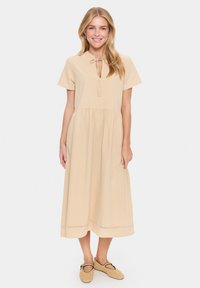 Rochie maxi din bumbac bej, cu mâneci scurte, decolteu legat și fustă băgată. Pur tată cu pantofi bej țesuti, cu un detaliu negru.