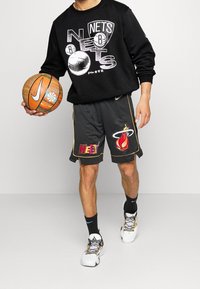 Felpa nera con logo "Nets", pantaloni corti da basket grigi con logo "Heat", in mano un pallone da basket, indossando calze sportive nere e scarpe bianche.