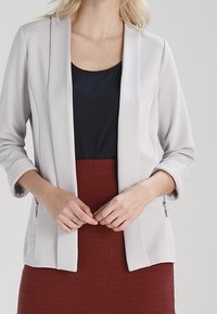 Blazer a coste grigio chiaro con apertura frontale, maniche a tre quarti e senza chiusure. Abbinato a un top blu navy e a una gonna texturizzata bordeaux.