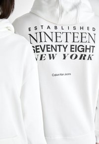 Bílá mikina s kapucí, která na zádech má velký černý grafický potisk s textem: "ZALOŽENO DEVADESÁT SEDM NEW YORK."
