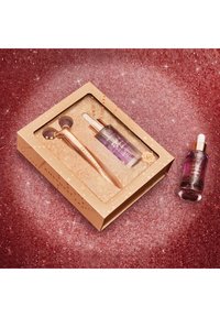 Zestaw do pielęgnacji skóry w kolorze różowego złota, zawierający wałek i butelkę z kroplomierzem. Przezroczysta szklana butelka z fioletową cieczą. Pudełko z błyszczącymi złotymi akcentami.