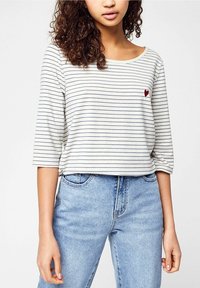 Randig, grå och vit långärmad skjorta med avslappnad passform och ett litet rött hjärtemblem på bröstet, tillsammans med ljusblå denimjeans.