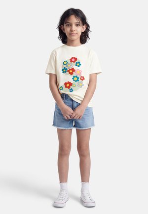 Enfant debout, les mains jointes, portant un t-shirt crème à motifs floraux colorés, un short en jean, des chaussettes blanches et des baskets blanches.