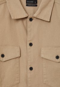 Camicia beige con bottoni, caratterizzata da un colletto, due tasche anteriori, bottoni neri e una vestibilità regolare. Realizzata in tessuto testurizzato con trame diagonali.