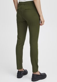 Olivnozeleni oprijeti hlače iz gladkega materiala z dvema zadnjima žepoma in srednjim pasom, v kombinaciji z črnimi robustnimi čevlji.