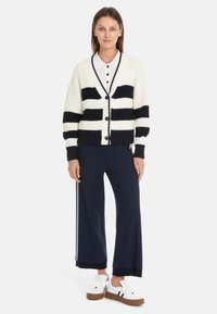 Strickjacke in Creme mit dunkelblauen Streifen, Knopfverschluss und gerippten Ärmeln, kombiniert mit dunkelblauen Hosen und weißen Sneakern mit braunen Sohlen.