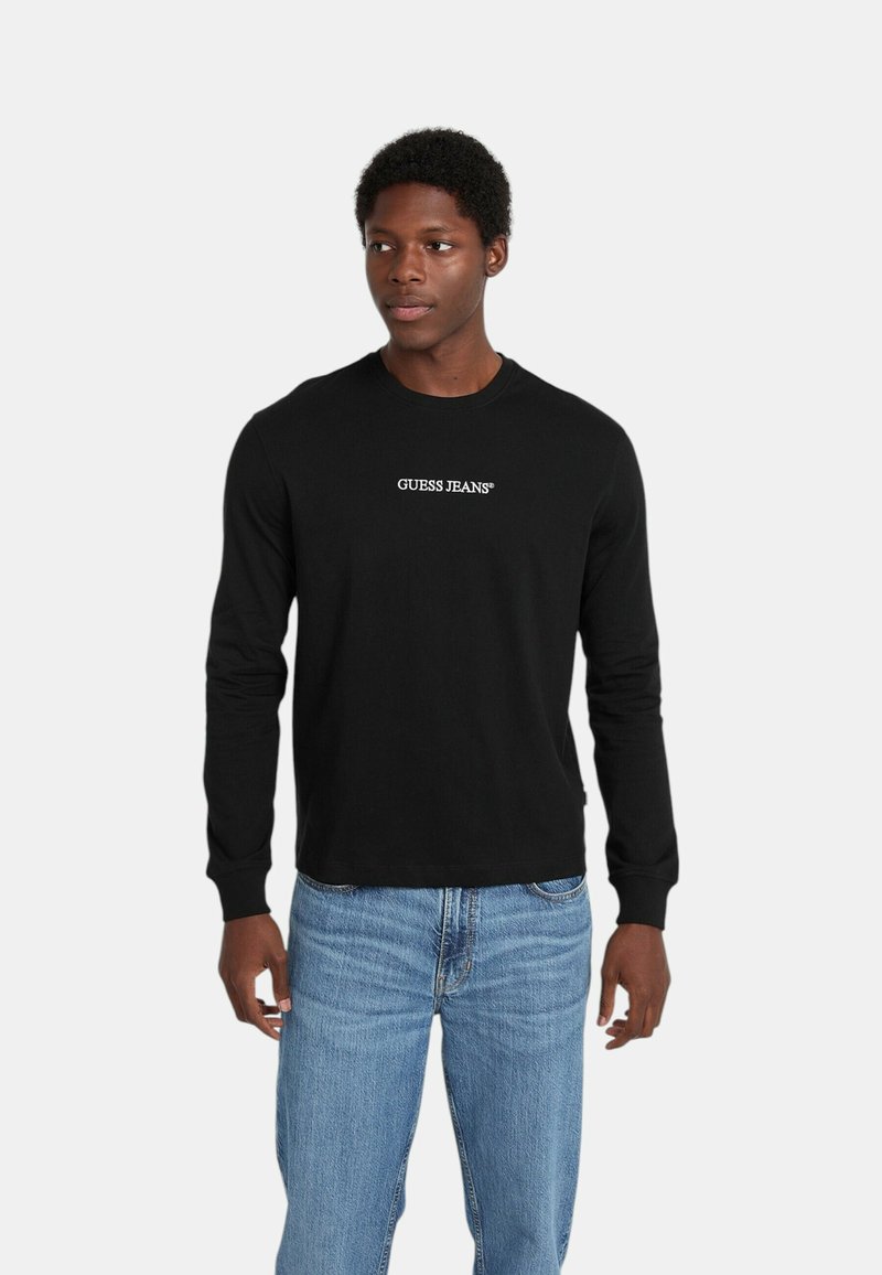 Maglietta nera a maniche lunghe con logo bianco "GUESS JEANS", abbinata a jeans di denim azzurri. Il tessuto sembra morbido e la vestibilità è rilassata.