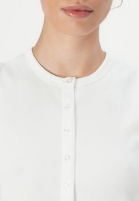 White ribbed top featuring a round neckline and six clear buttons down the front. Soft texture, fitted shape, and plain design.