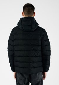 Giacca a vento nera con cappuccio, design a righe verticali e texture liscia, indossata sopra pantaloni in denim neri.