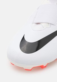 Scarpa da calcio bianca con il simbolo Nike nero, tomaia testurizzata e dettagli rosa brillante sulla suola. Presenta una fascia in velcro e un design a coste.