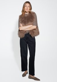 Gilet en fausse fourrure marron superposé à un haut en tricot marron clair, associé à un jean foncé et des chaussures slip-on taupe ; palette de couleurs neutres et texture douce.