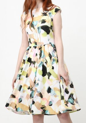 Ärmelloses weißes Kleid mit abstraktem Blumenmuster in Pastellrosa, Blau, Gelb, Schwarz und Grün, getragen von einer Frau mit roten Haaren.