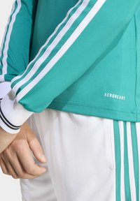 AeroReady sportinis viršus melsvai žalias su baltais dryžiais ant rankovių, su raukšlėtu rankogaliu ir logotipo detalėmis hem'e. Matomos baltos kelnės.