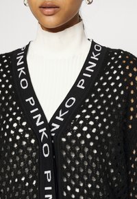 Svart, öppet stickad cardigan med ett kontrasterande vitt logoband längs med halsringningen, lager-på-lager över en vit ribbad polotröja.