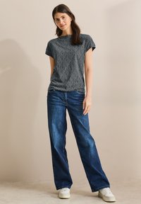 Šedé tričko s krátkým rukávem a vzorem, doplněné volnými modrými džíny a bílými teniskami, na neutrálním pozadí.