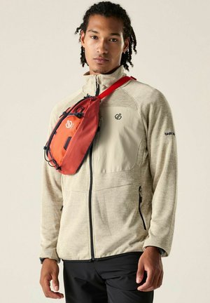 Jonge man met een beige jas met ritssluiting en een rode crossbodytas tegen een effen lichte achtergrond.