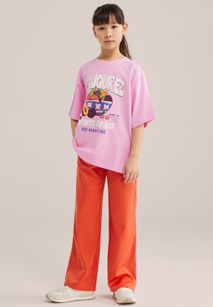 Ragazza giovane con capelli scuri raccolti in una coda di cavallo, che indossa una maglietta rosa oversize con stampa di frutta e pantaloni arancioni a gamba larga, abbinati a sneakers bianche.