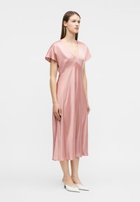 Γυναίκα που στέκεται με ροζ μίντι φόρεμα από σατινέ ύφασμα με κοντά μανίκια και V-λαιμόκοψη, φοράει λευκά μυτερά παπούτσια με χαμηλό τακούνι, σε λευκό φόντο.