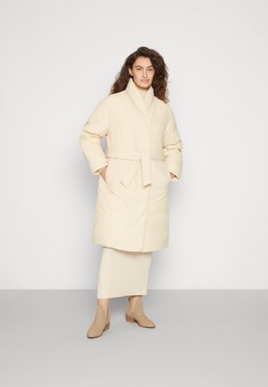 Manteau rembourré beige avec une ceinture, grands revers et manches longues, présentant une longueur mi-cuisse et une texture douce. Porté par-dessus une tenue assortie.