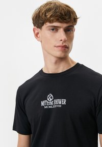 Sort bomulds T-shirt med hvid grafisk tekst "METEOR SHOWER" og "MCMLXXXVIII" på brystet. Rund hals og korte ærmer.