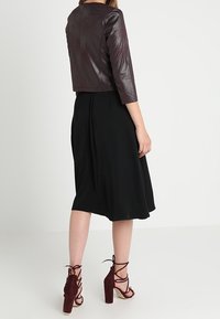 Veste courte en cuir bordeaux, jupe noire tombant sous le genou, et talons lacés bordeaux à talons carrés visibles de dos.