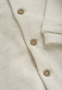 Beige gestrickte Strickjacke mit braunen Vierlochknöpfen und einem gerippten Bündchen am Ärmel, in Nahaufnahme gezeigt.