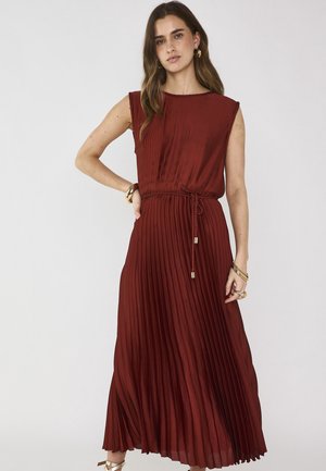 Giovane donna con capelli lunghi in abito senza maniche plissettato rosso scuro, legato in vita, che indossa bracciali, anelli e orecchini a cerchio dorati.