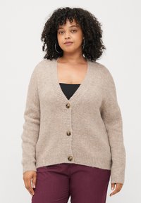 Beige pletený cardigan s véčkovým výstřihem, žebrovanými manžetami a třemi knoflíky vpředu, v kombinaci s tmavě kaštanovými kalhotami. Měkká textura.