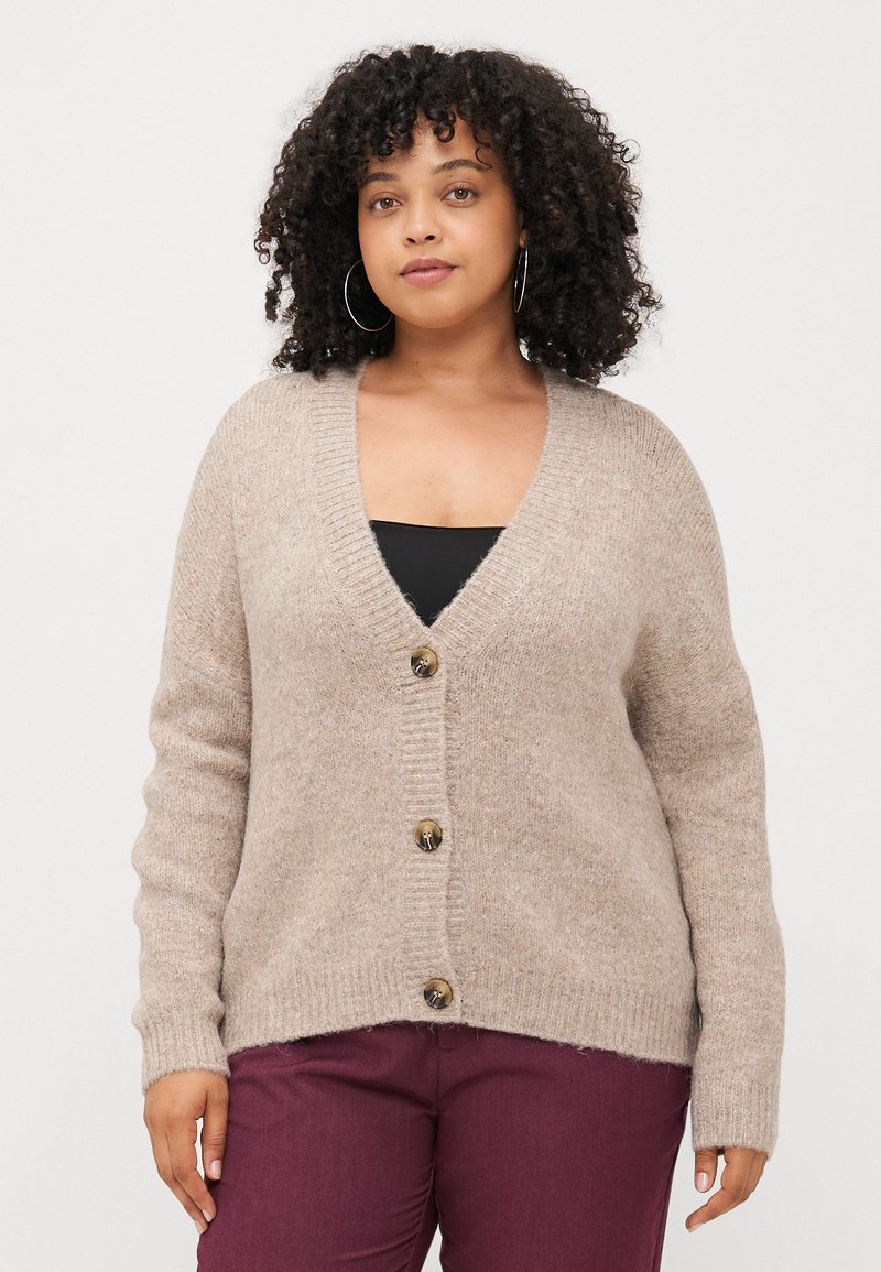 Beige pletený cardigan s véčkovým výstřihem, žebrovanými manžetami a třemi knoflíky vpředu, v kombinaci s tmavě kaštanovými kalhotami. Měkká textura.
