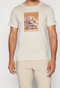 Lys beige kortærmet t-shirt med en bjerggrafik og teksten "Columbia Sportswear Co." på brystet, båret med lys beige bukser.