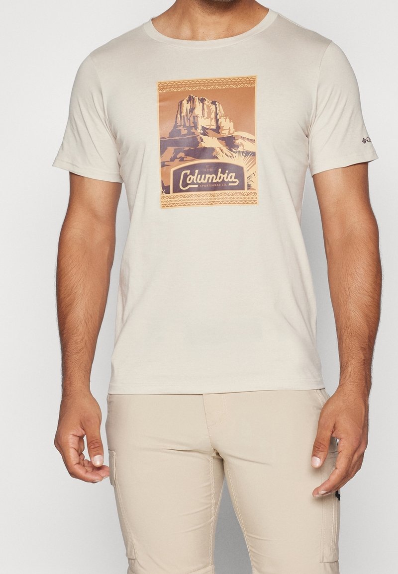 Lys beige kortærmet t-shirt med en bjerggrafik og teksten "Columbia Sportswear Co." på brystet, båret med lys beige bukser.